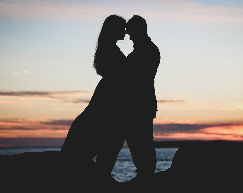 Mann und Frau stehen sich gegenüber, umarmen sich, Stirn an Stirn, beide stehen auf Felsen vor dem Meer bei Sonnenuntergang